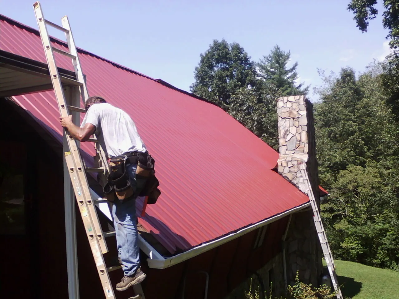 Roofer on a ladder beside a red metal roof and stone chimney — Soffit & Fascia Repair by Roofing Brothers in Oak Harbor